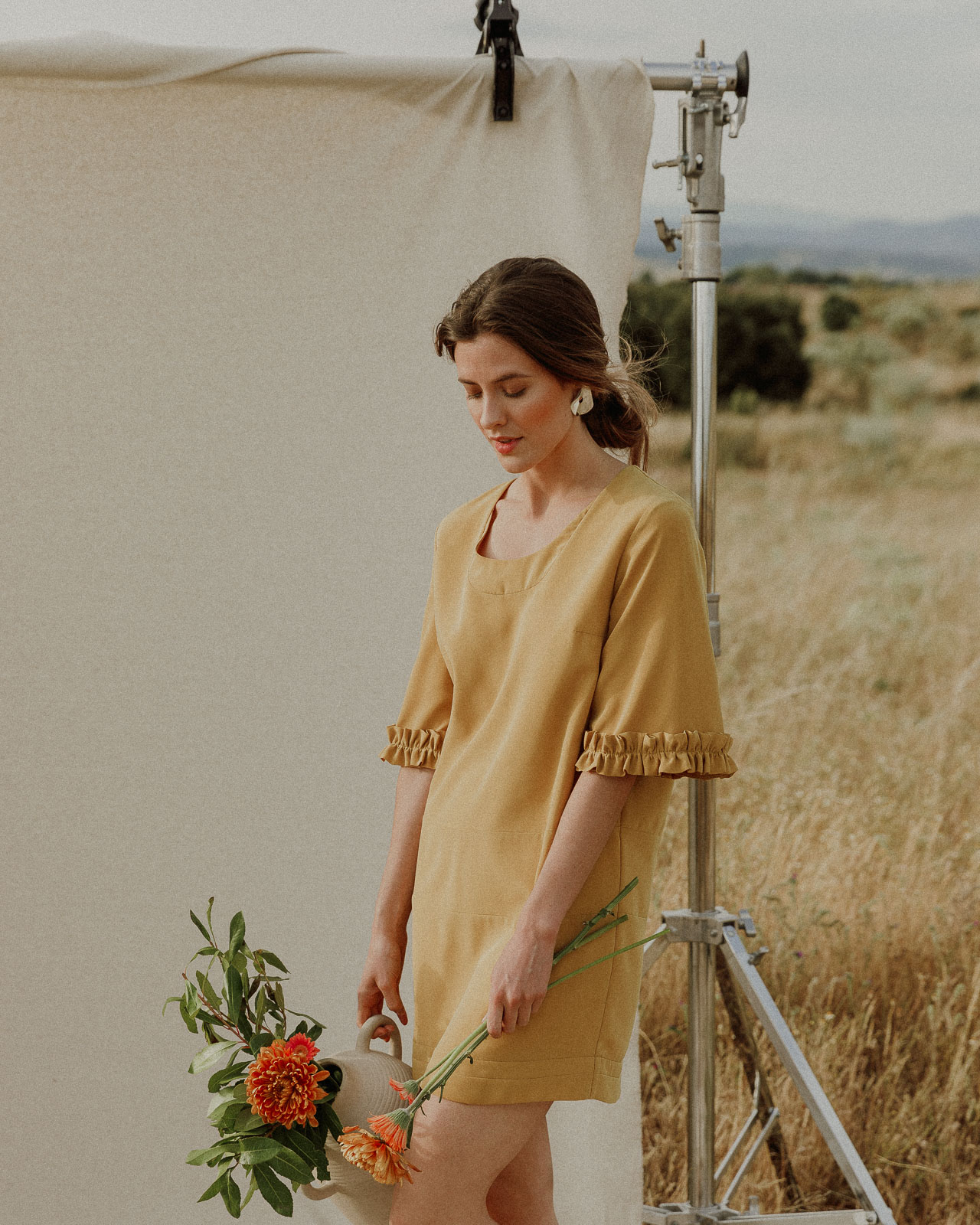 Mujer con vestido mostaza en un campo seco, sosteniendo una jarra con flores